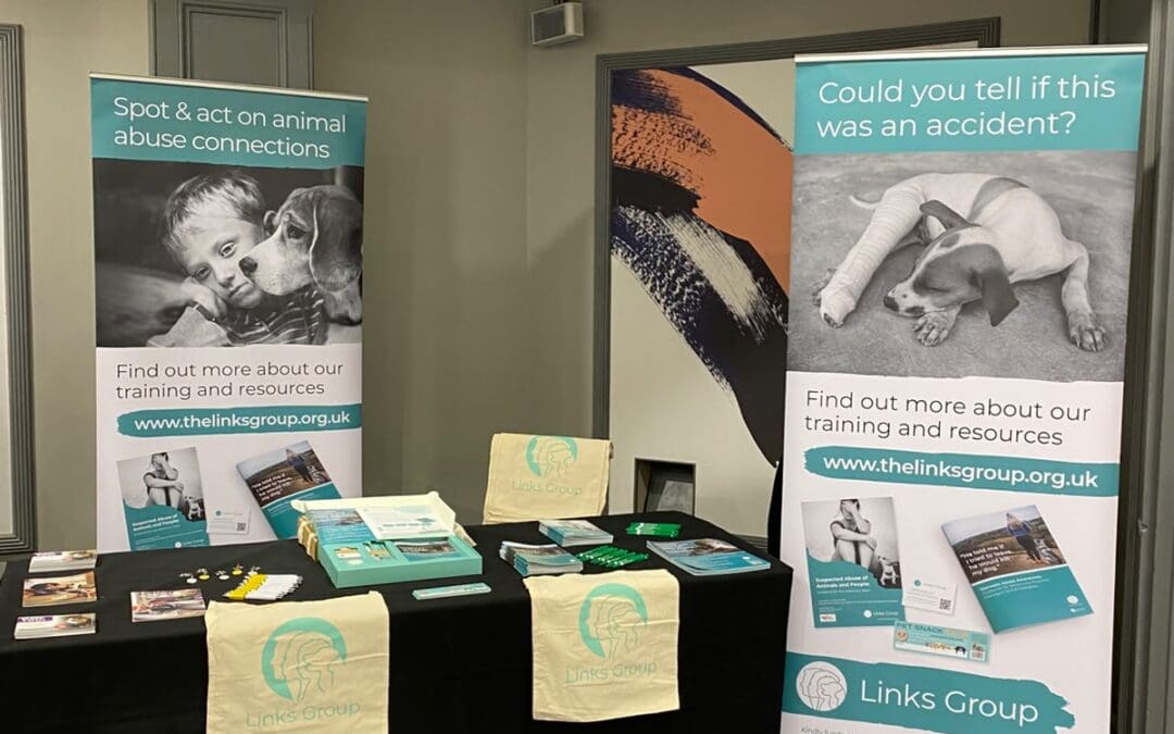 Exhibition table with brochures, tote bags, and two banners about animal abuse awareness from The Links Group, featuring images of a dog and a cat. Displayed at BVRA Congress 2026, the stand highlights vital information about preventing animal abuse.