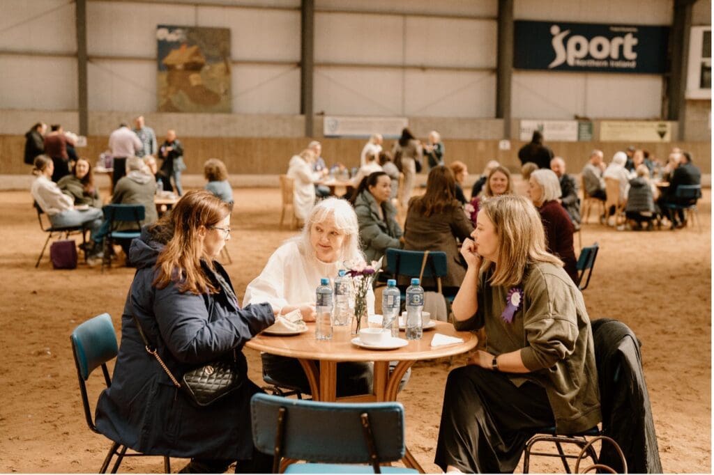 A group of people sit at round tables in a large indoor venue during a tea break at Causeway Coast & Glens RDA - Coleraine, chatting and drinking water in a relaxed, social atmosphere with banners as decoration.