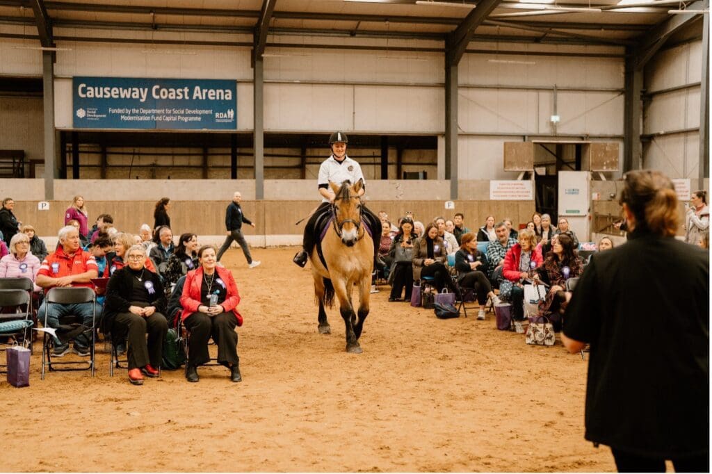 A person rides a horse down an aisle in the Causeway RDA - Indoor Arena, with seated spectators on both sides and a banner reading Causeway Coast Arena in the background.