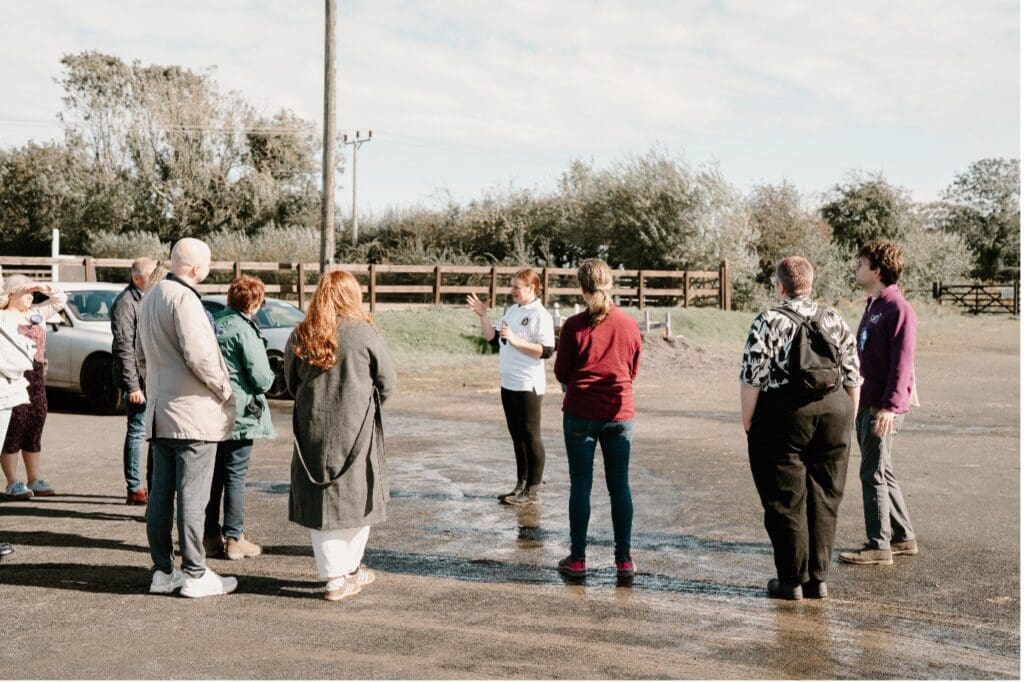 A group of people stands in a car park at RDA - Coleraine, listening to a person speak. Trees, a fence, and cars are visible in the background under a partly cloudy sky.