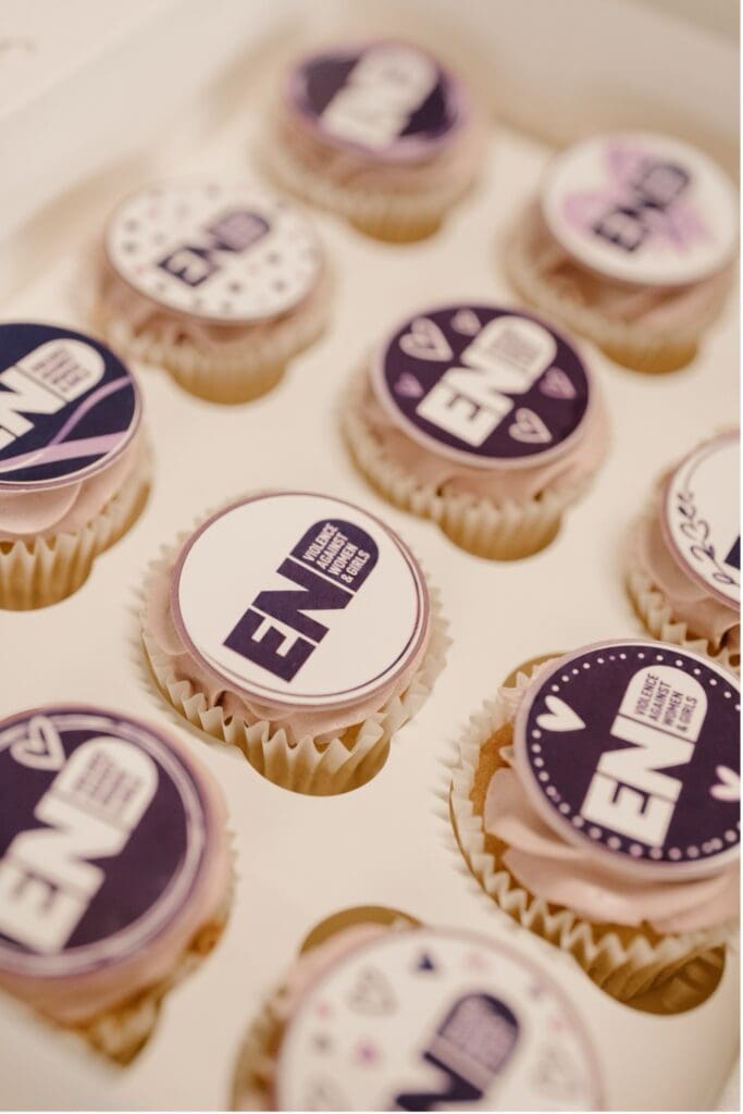 A box of cupcakes with purple and white icing, each topped with an “END” logo and heart or dot designs. These cupcake toppers by Lesley at Twiggybee support End Violence against Women—“Every Needs Food.”.