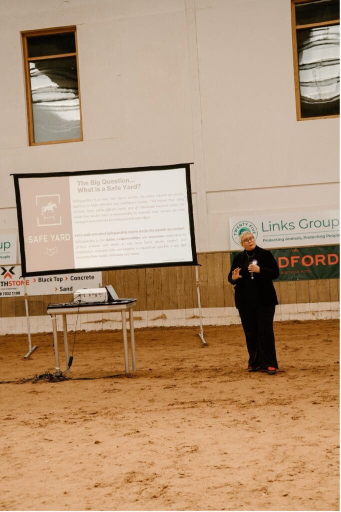 A person in a suit stands in an indoor arena, giving a presentation with a microphone. Speaker Marcella Leonard discusses the “Safe Yard” as a projector screen displays info. A table holds a laptop and cables, and signs are on the walls.