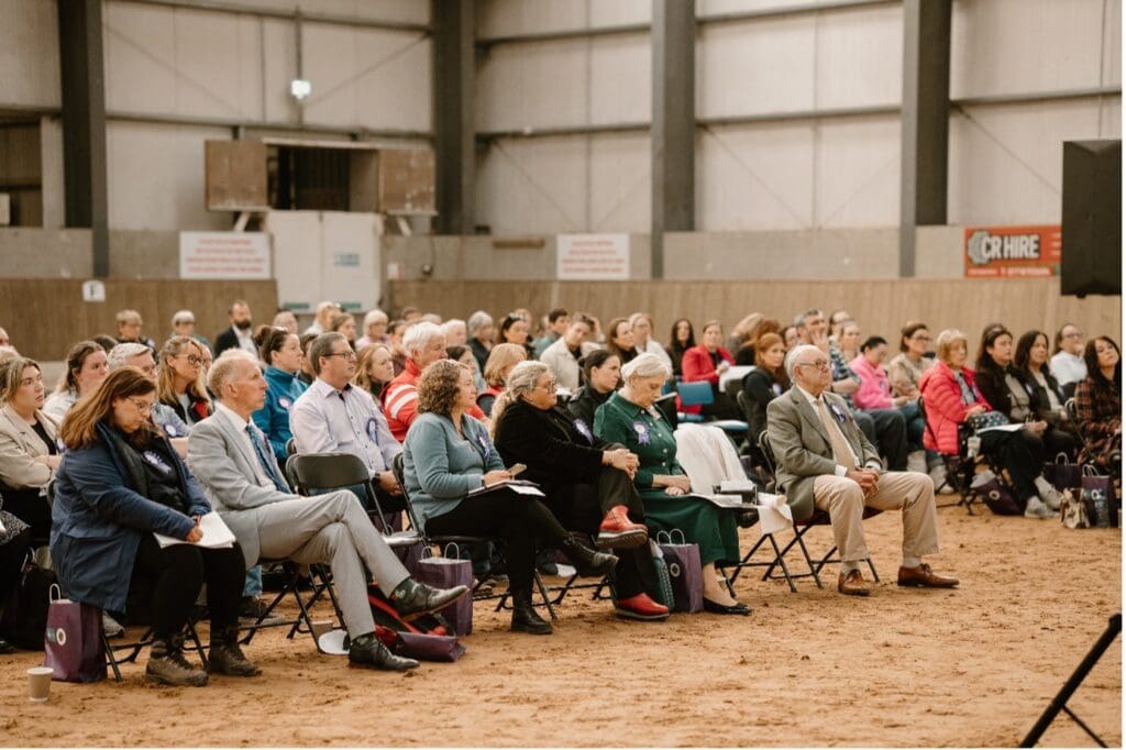 Delegates at Causeway Coast & Glens RDA: The Link – Connecting Cruelty sit on folding chairs in an indoor arena, attentively watching the event. Most are middle-aged or older, engaged and taking notes amid industrial walls and dirt flooring.
