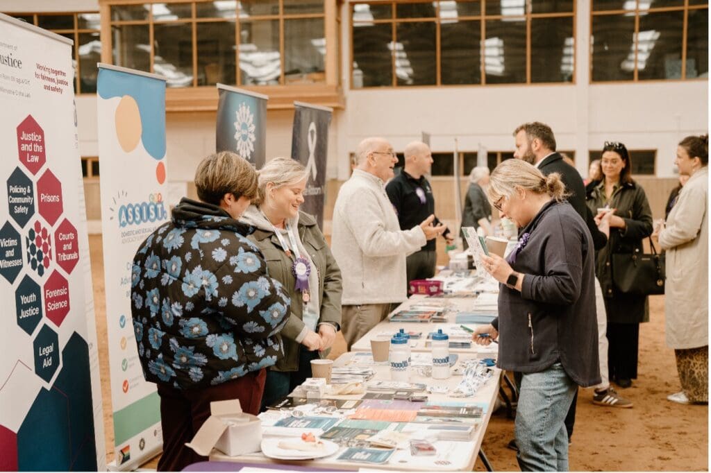 People interact at an indoor information fair with Trade Stands at The RDA - Coleraine, visiting tables with brochures, banners, and merchandise. Attendees wearing lanyards chat and browse materials in a warm, engaged atmosphere.